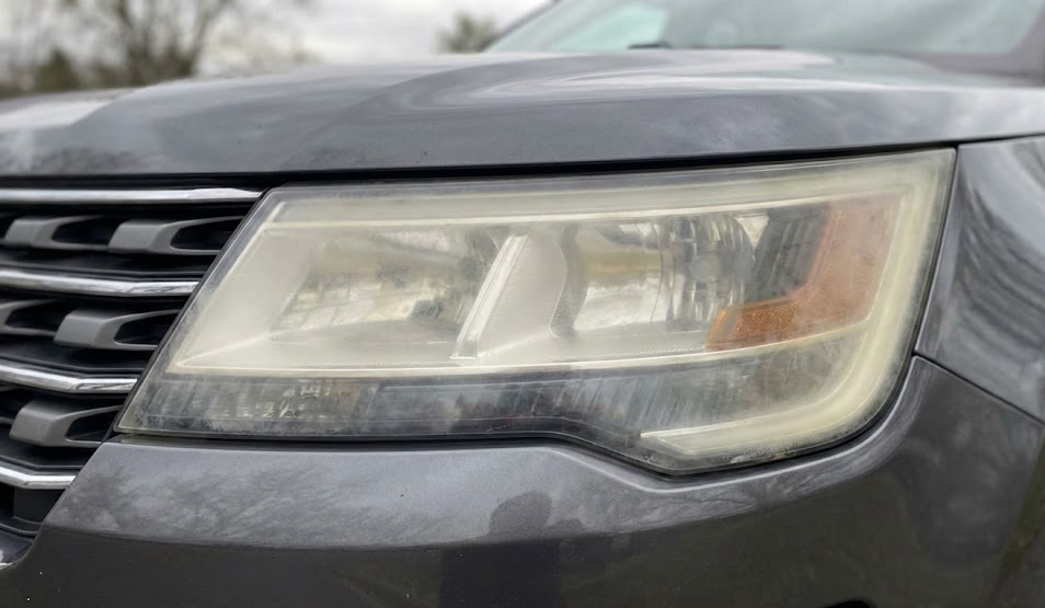 close up of cloudy oxidized headlight lens on gray SUV, hazy yellowed plastic reducing light output, example of dim headlights before restoration, Athens Ford headlight restoration service Athens GA, visibility problems for Ford Explorer and Escape drivers, Georgia UV sun damage to headlight lenses, need for professional cleaning and UV protective coating, dealership service center demonstrating before and after clarity, safety reminder for nighttime driving during deer season, local Ford dealer maintenance photo