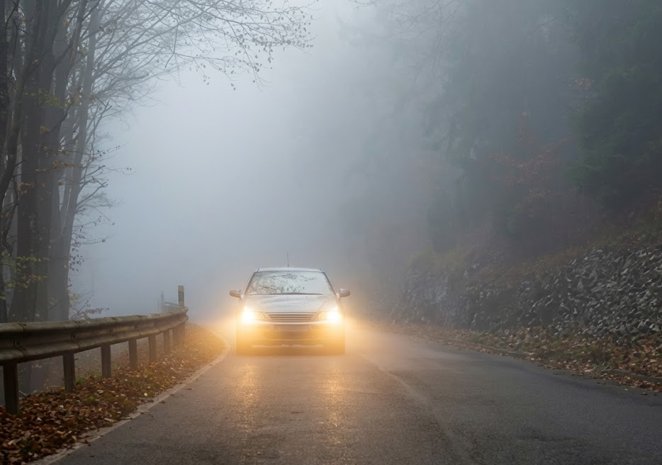 Foggy mountain road at dusk near Colorado Springs, car headlights struggling through low visibility, dangerous November night driving on I-25 and Powers Boulevard, deer and elk collision risk in the Pikes Peak region, Colorado winter driving conditions with poor headlight performance, cloudy headlights reducing visibility, Nissan driver needs professional headlight restoration and winter visibility inspection, South Colorado Springs Nissan service center, Colorado Springs CO