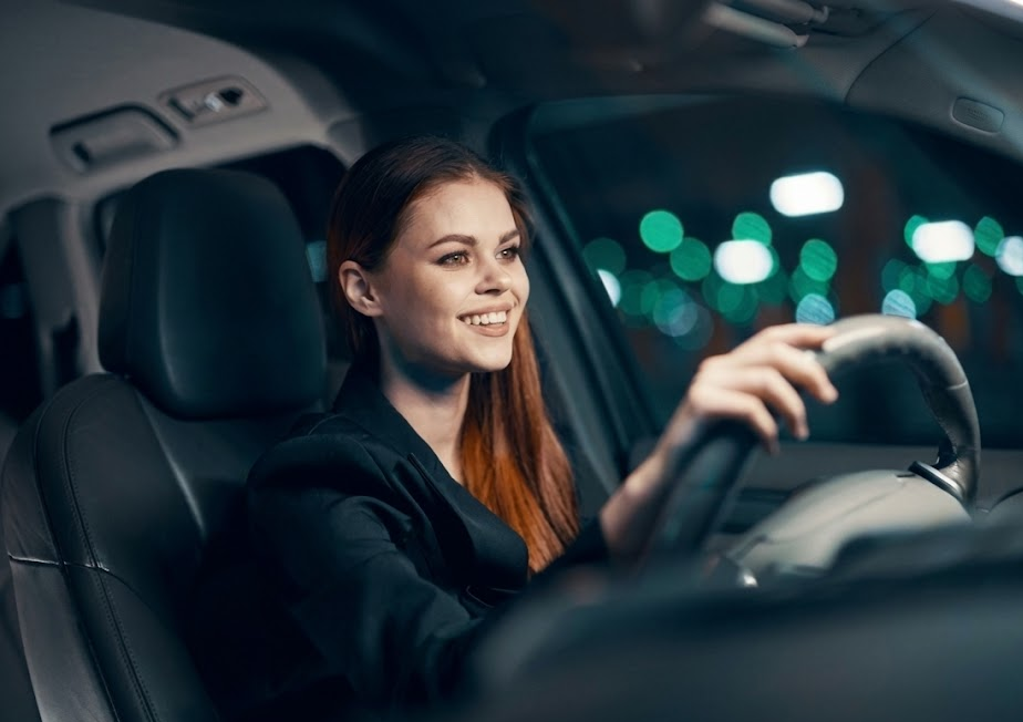 Smiling young woman confidently driving Nissan SUV at night, clear windshield and bright headlights, safe winter visibility after professional service, Colorado Springs city lights in soft blur, driver relaxed after headlight restoration and new beam-style wiper blades, Nissan winter safety inspection completed, South Colorado Springs Nissan service customer, confident nighttime driving in Colorado