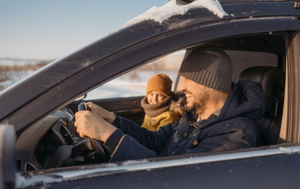 Happy father and child riding in warm SUV on sunny winter day, smiling family enjoying safe winter drive after installing snow tires, snow visible on car roof and in background fields, bundled up in hats and coats for cold Colorado Springs weather, confident driver holding steering wheel with excellent traction and control, family safety thanks to dedicated winter tires and brake inspection, Woodmen Nissan Service customer satisfaction, secure winter driving near Pikes Peak and Colorado Springs neighborhoods