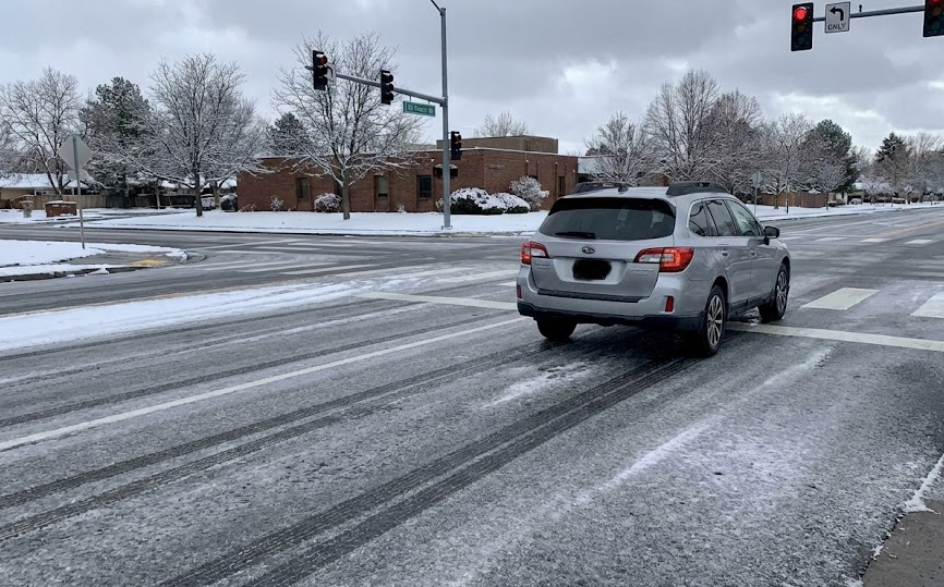 Car braking at icy urban intersection with red traffic light, snowy Colorado Springs neighborhood streets, slick black ice on road surface showing long tire tracks, dangerous winter stopping distance near busy crosswalk, Colorado winter commute on streets like Woodmen Road and Academy Boulevard, driver needing reliable winter tires and good brakes, cold gray sky and light snow on trees and sidewalks, Woodmen Nissan Service winter safety inspection and brake check for Nissan vehicles