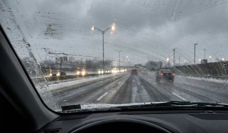 Driver view through streaked windshield in winter traffic, mag chloride film smeared across glass, headlights and taillights blurred by greasy road spray, Colorado Springs highway in snow and slush, standard wiper blades failing to clear de-icer, poor visibility during storm, need for beam-style winter wiper blades and quality washer fluid, Nissan winter visibility service, South Colorado Springs Nissan on S. Academy Blvd