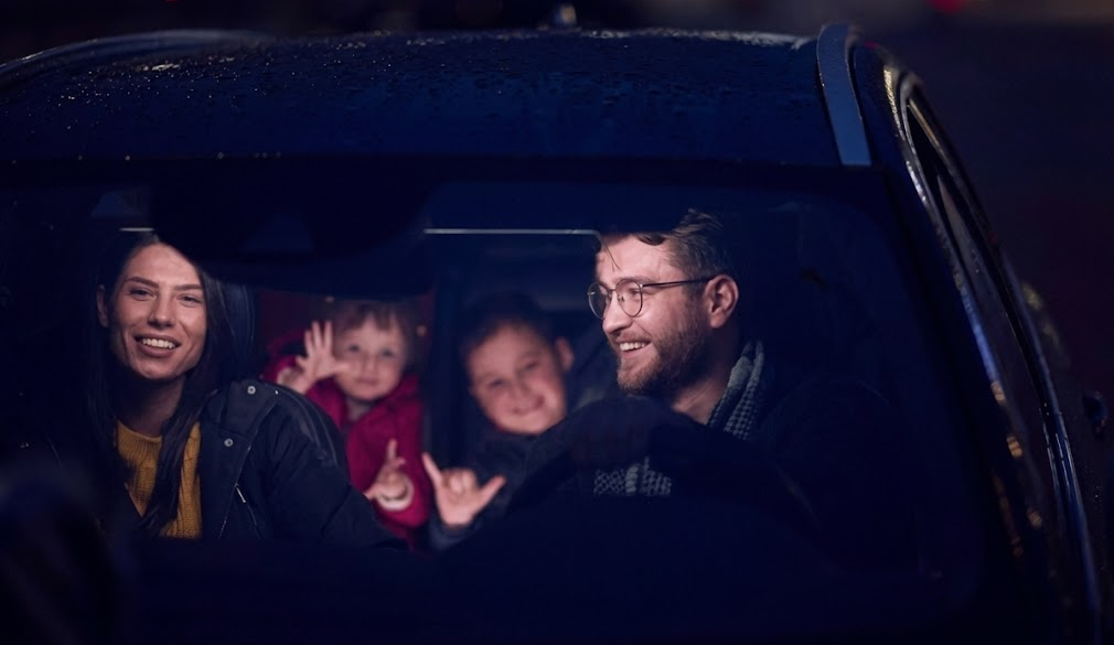 smiling family riding together in car at night, confident dad driving with kids and mom in front seat, safe Nissan SUV on wet road, headlights reflecting after brake inspection, Cookeville TN family road trip, peace of mind after Nissan brake service, Highway 111 holiday travel safety, Putnam County Nissan dealer, family protected by reliable OEM brakes, stress free deer season driving around Cookeville