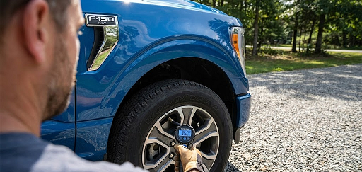 driver checking tire pressure on blue Ford F-150 with digital gauge, cold front tire pressure drop in Palm Bay Florida, Ford truck tire maintenance Brevard County, TPMS light prevention, Ford F-150 tire inspection in gravel driveway, Palm Bay Ford Service tire check, proper PSI for Ford pickup, Florida cold weather tire care, dealership tire service near Malabar Road