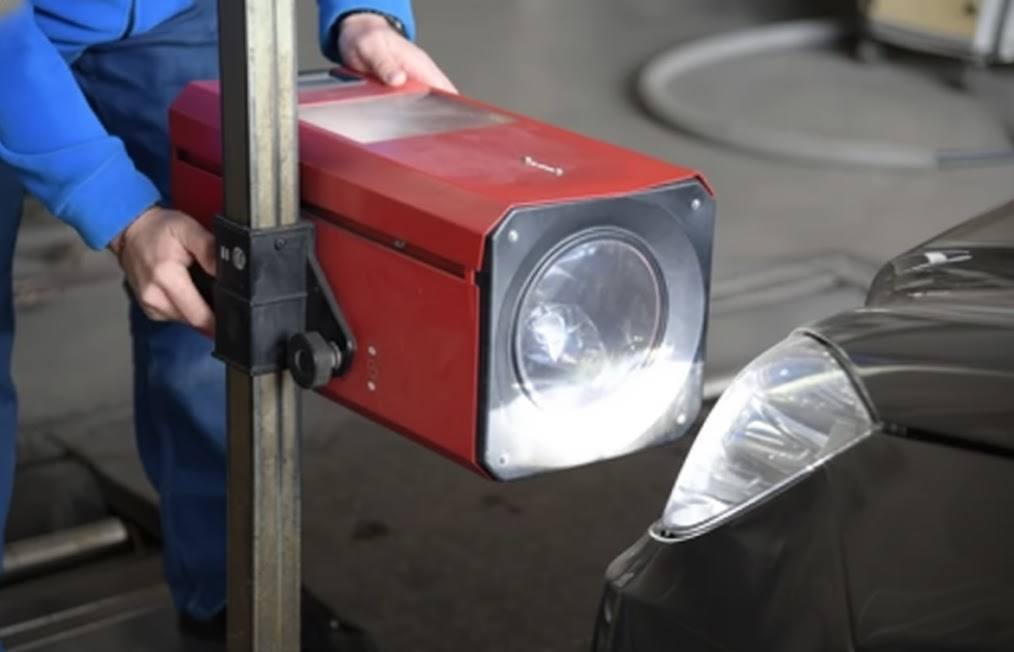 auto technician using professional headlight aiming machine, checking beam pattern in service bay, Scott Clark Nissan Service headlight alignment, Charlotte NC visibility inspection, adjusting low beam aim for safe night driving, Nissan sedan front headlamp close up, fall driving safety on I-77 and I-485, correcting dim headlights and glare, factory-trained technician using alignment equipment, headlight restoration and aiming service in Charlotte