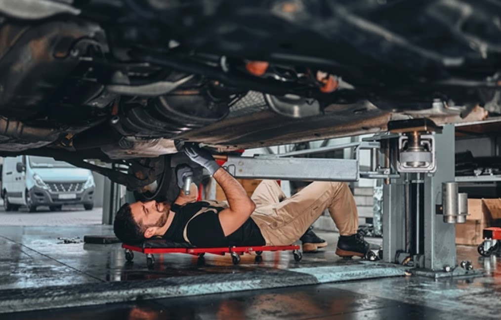 Certified technician lying on creeper under vehicle in service bay, inspecting transmission and driveline components, professional Nissan CVT maintenance at Woodmen Nissan Service Colorado Springs, severe service CVT fluid exchange, factory-trained mechanic checking for leaks and wear, dealership lift equipment, transmission protection before mountain driving trip
